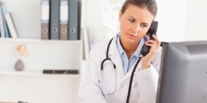 Foto einer Ärztin im Büro mit Stethoskop um den Hals telefoniert und sieht auf einen Computerbildschirm